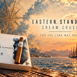 Caldwell Eastern Standard Cream Crush cigar and box against a nautical sunset harbor with vintage compass artwork.